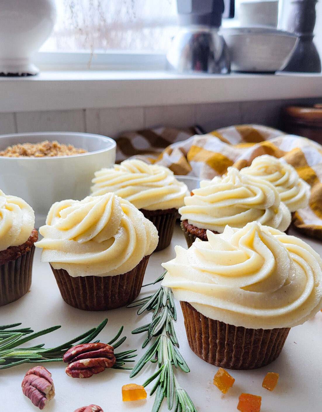 Freshly frosted Best Carrot Cake Cupcakes with Ginger Cream Cheese Frosting are on the counter, waiting to be garnished with herbs and chopped dried apricots.