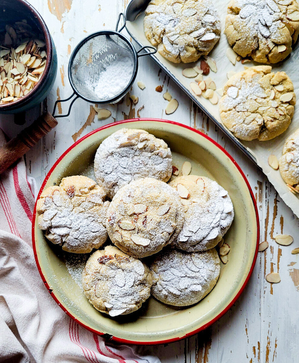 A platter filled with Almond Croissant Cookies on the table, with sliced almonds and icing sugar to the side.