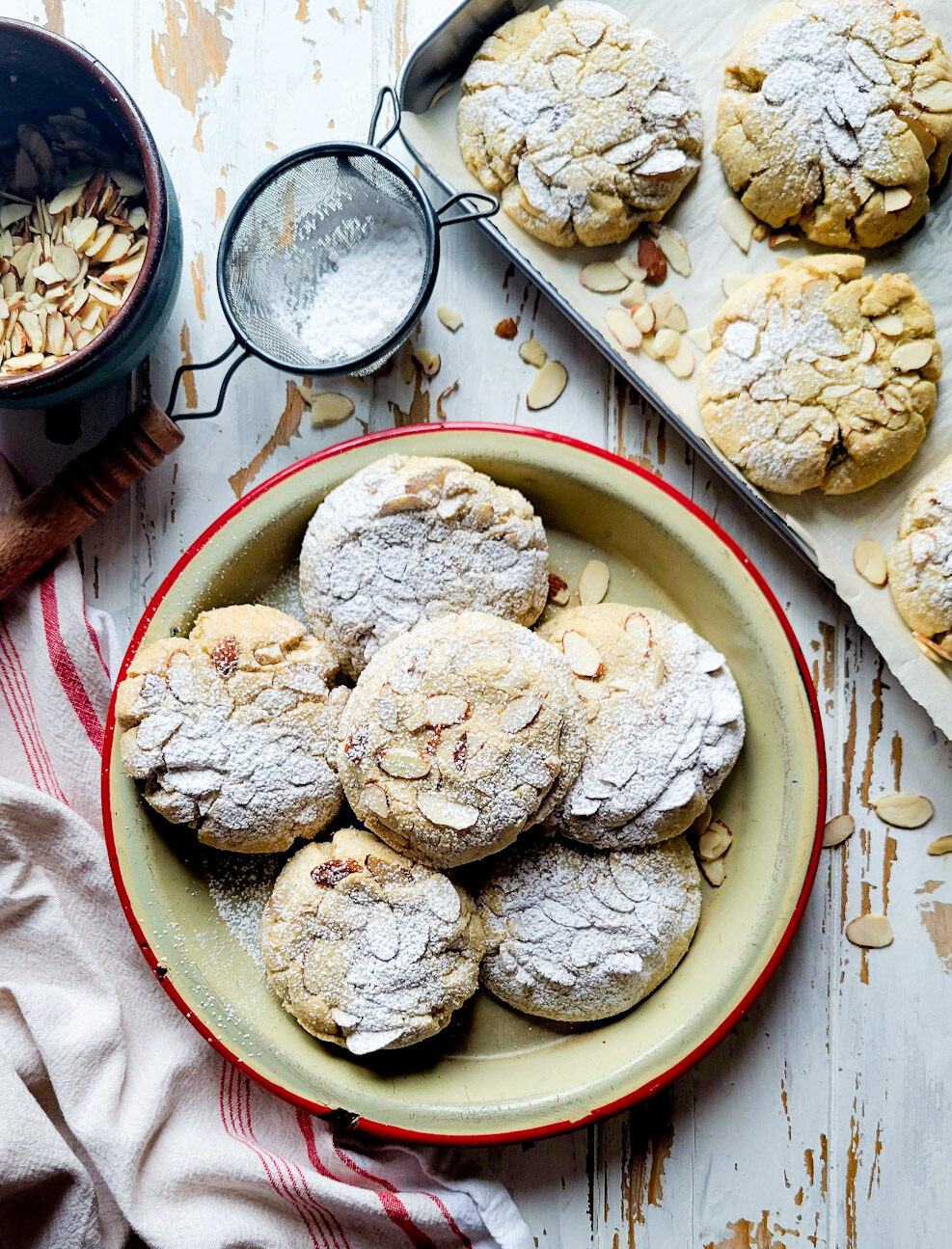 A platter filled with Almond Croissant Cookies on the table, with sliced almonds and icing sugar to the side.