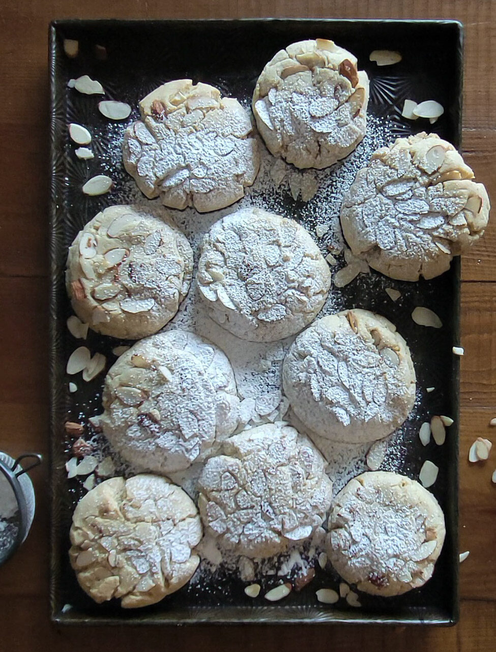 Almond Croissant Cookies on the baking sheet, dusted with icing sugar, wtih extra almond slices scattered about.