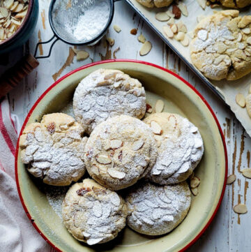 A platter of Almond Croissant Cookies dusted with icing sugar on the table, with more cookies on the baking sheet to the side.