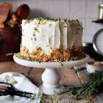 The Best Carrot Cake with Ginger Cream Cheese Frosting sits on stand with fresh thyme sprigs to the side.