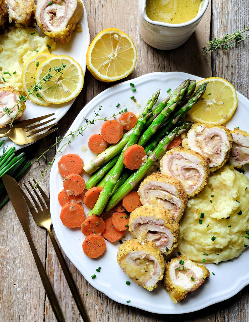 Two plates with Chicken Cordon Bleu sliced and served with mashed potatoes, asparagus and carrots. A container of lemon butter sauce is to the side.