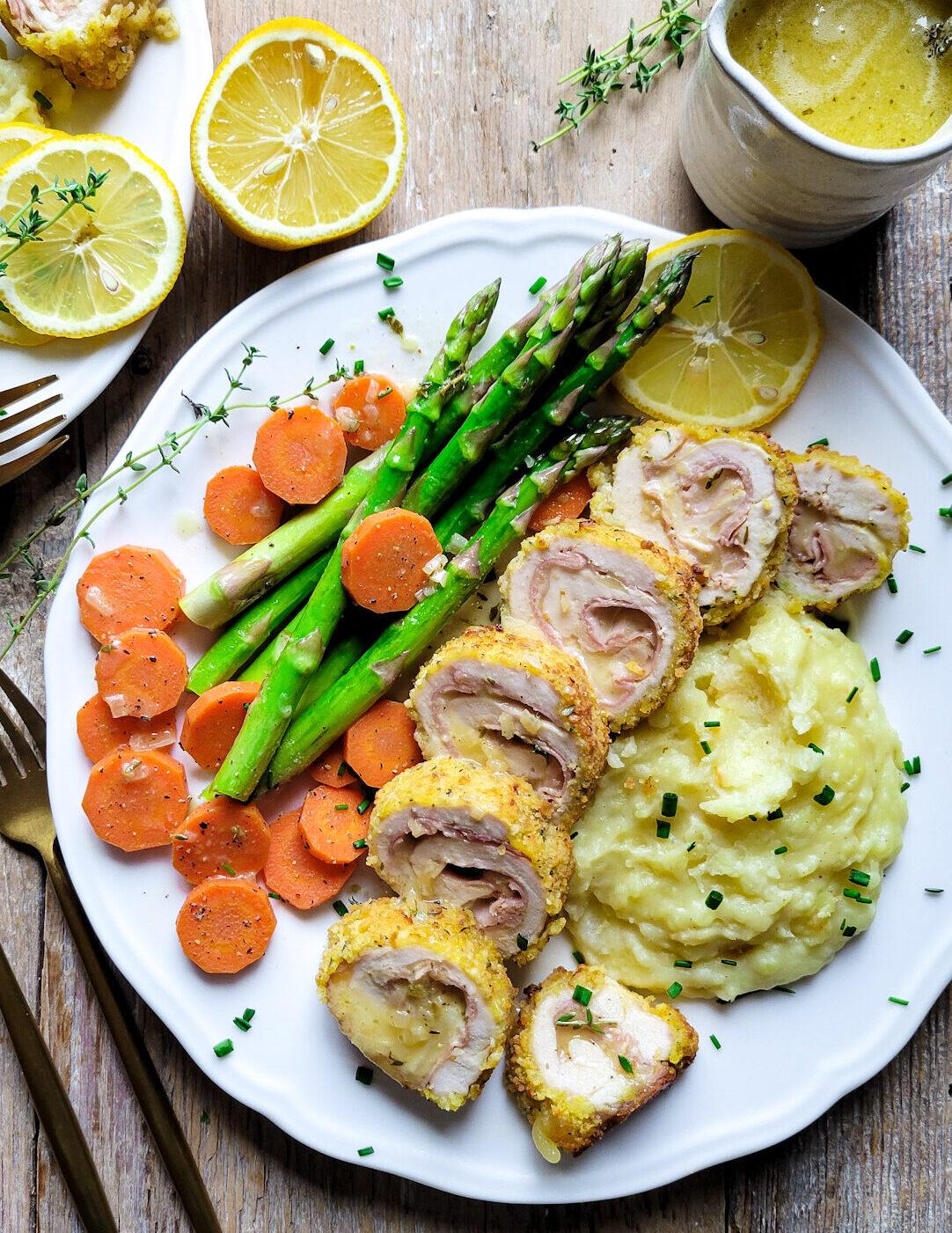 Two plates with Chicken Cordon Bleu sliced and served with mashed potatoes, asparagus and carrots. A container of lemon butter sauce is to the side.