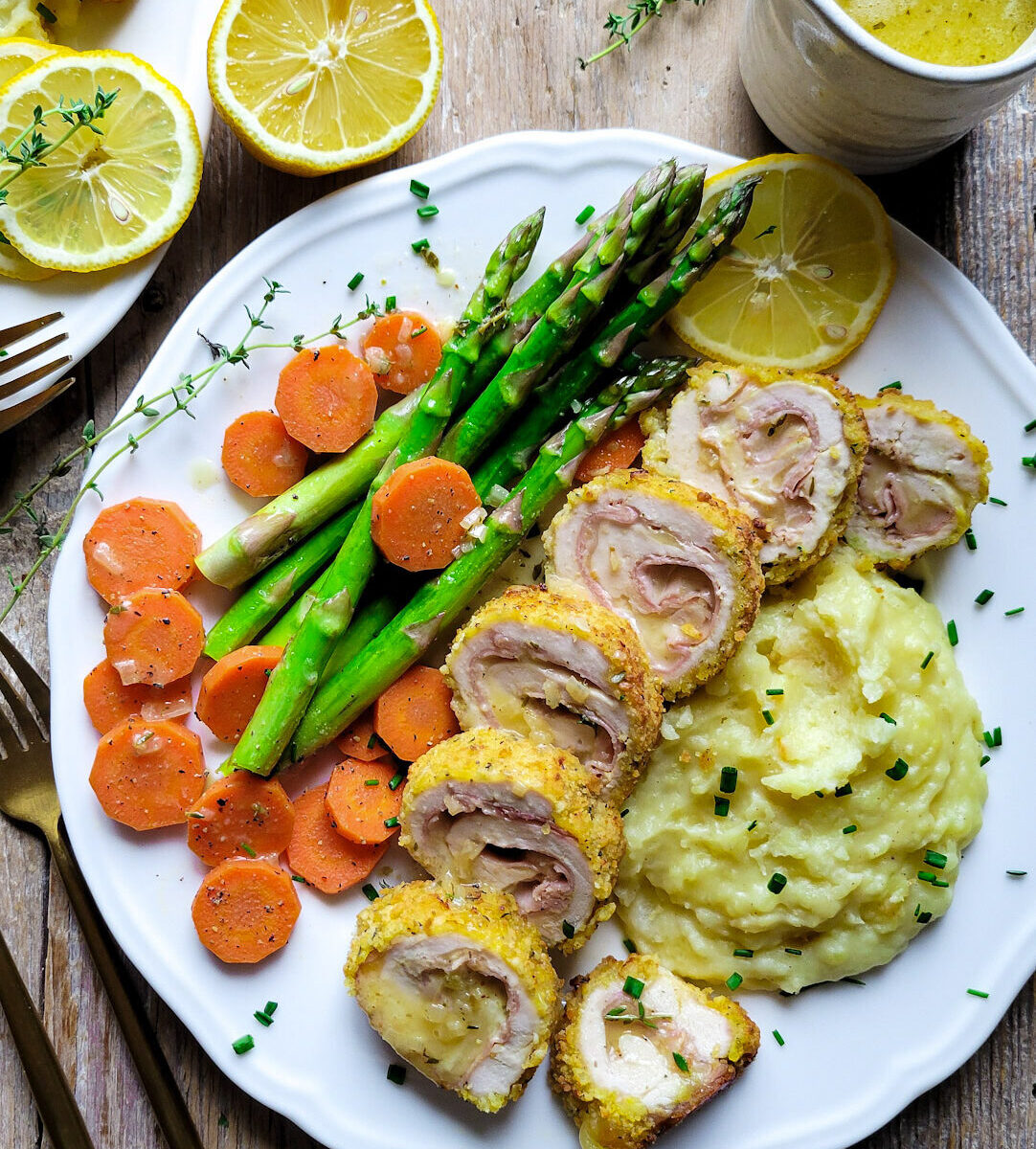 A plate set with Chicken Cordon Bleu, mashed potatoes, carrots and asparagus. Lemon Butter Sauce is to the side.