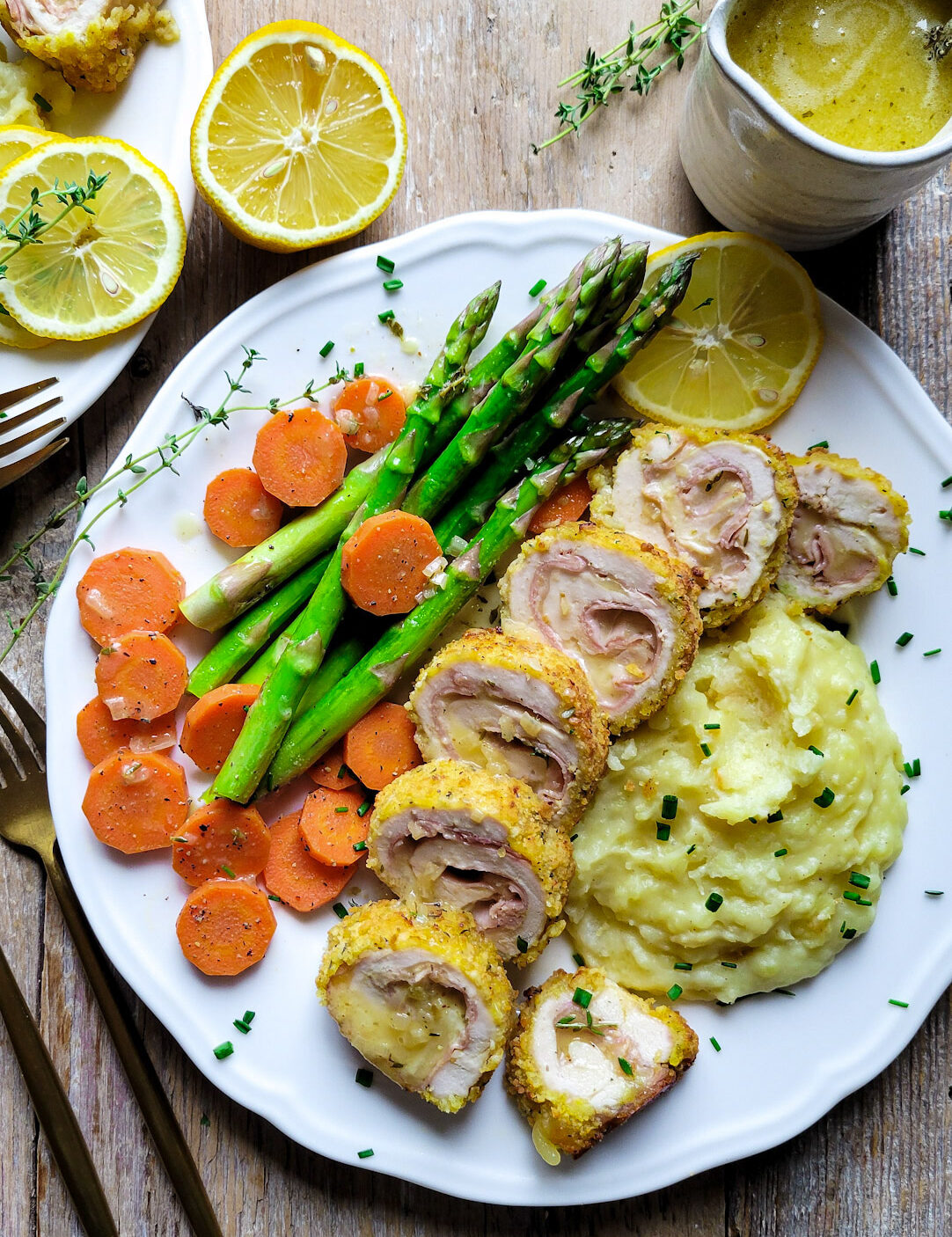 A plate set with Chicken Cordon Bleu, mashed potatoes, carrots and asparagus. Lemon Butter Sauce is to the side.
