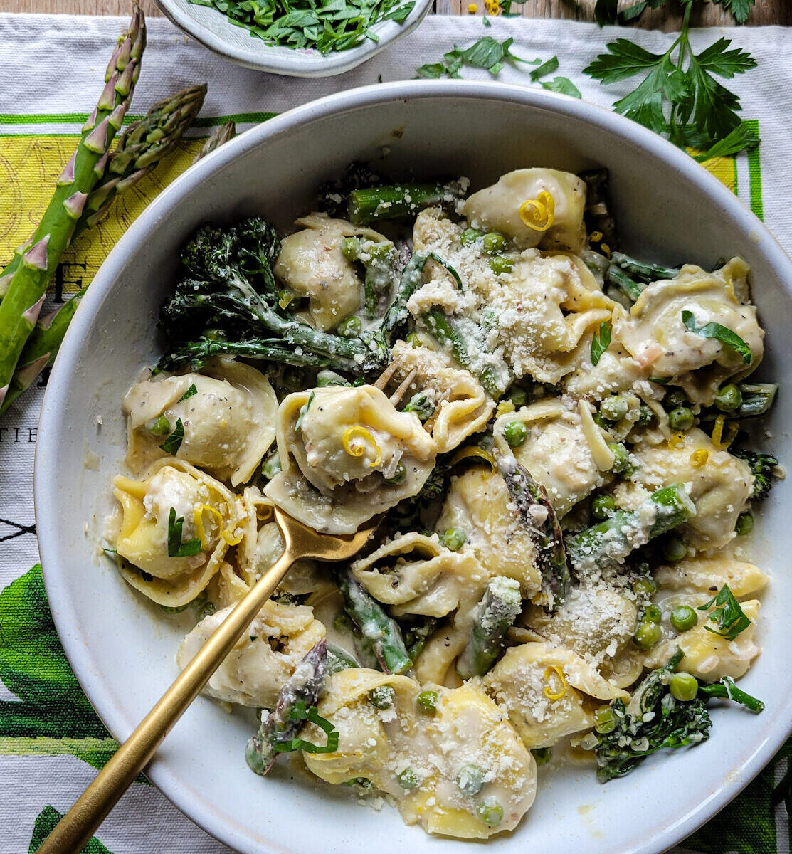 A bowl of Lemon Cream Pasta with Spring Vegetables with a fork holding some pasta is on the table, with fresh asparagus and parsley in the background.