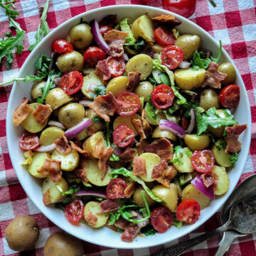 A bowl of succulent BLT Potato Salad, with mini potatoes, bacon, arugula, grape tomatoes, and red onion sits on a red checkered cloth with serving utensils to the side.