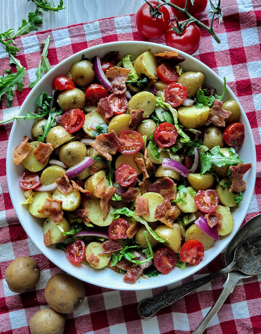 A bowl of succulent BLT Potato Salad, with mini potatoes, bacon, arugula, grape tomatoes, and red onion sits on a red checkered cloth with serving utensils to the side.