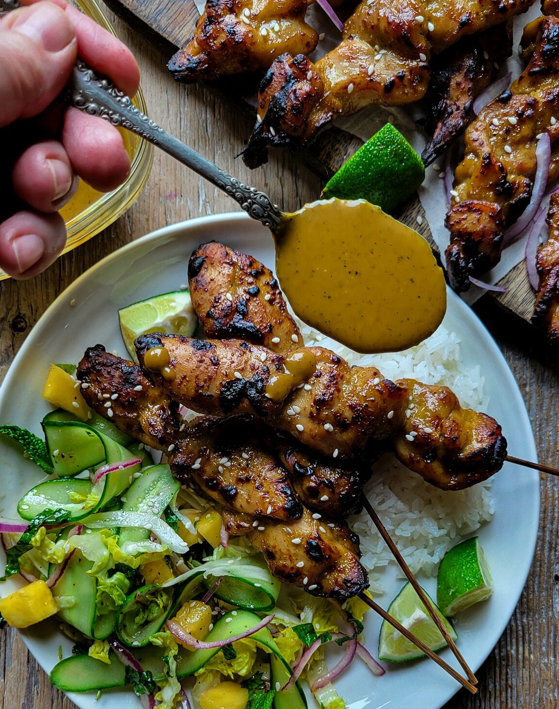 A spoon holding spicy Peanut sauce is being drizzled over Chicken Satay skewers on a plate with rice and tropical salad.