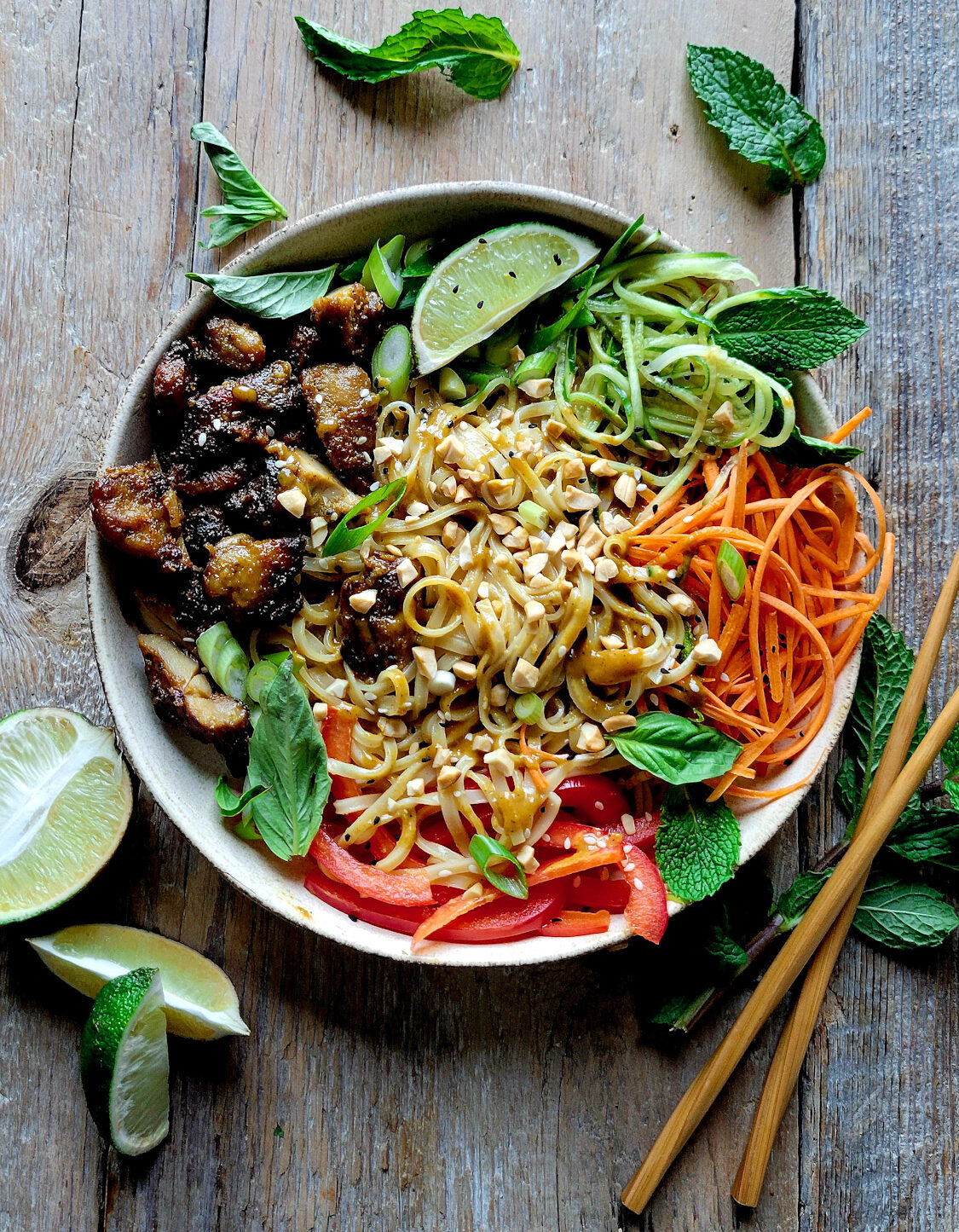 A Bowl filled with Chicken Satay, Peanut Noodles, and vegetables to create a flavour packed Satay Noodle Bowl in on the table, with mint leaves and limes to the side.