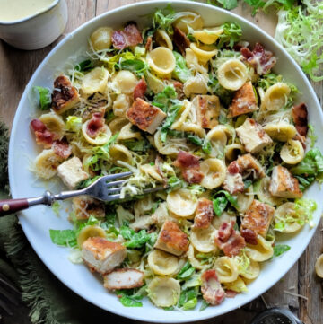 A bowl of Caesar Pasta Salad with Parmesan Crusted Chicken Croutons is surrounded by lettuce, cheese and creamy dressing.