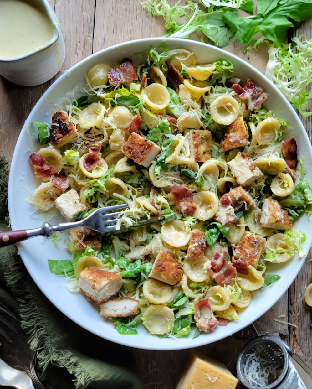 A bowl of Caesar Pasta Salad with Parmesan Crusted Chicken Croutons is surrounded by lettuce, cheese and creamy dressing.