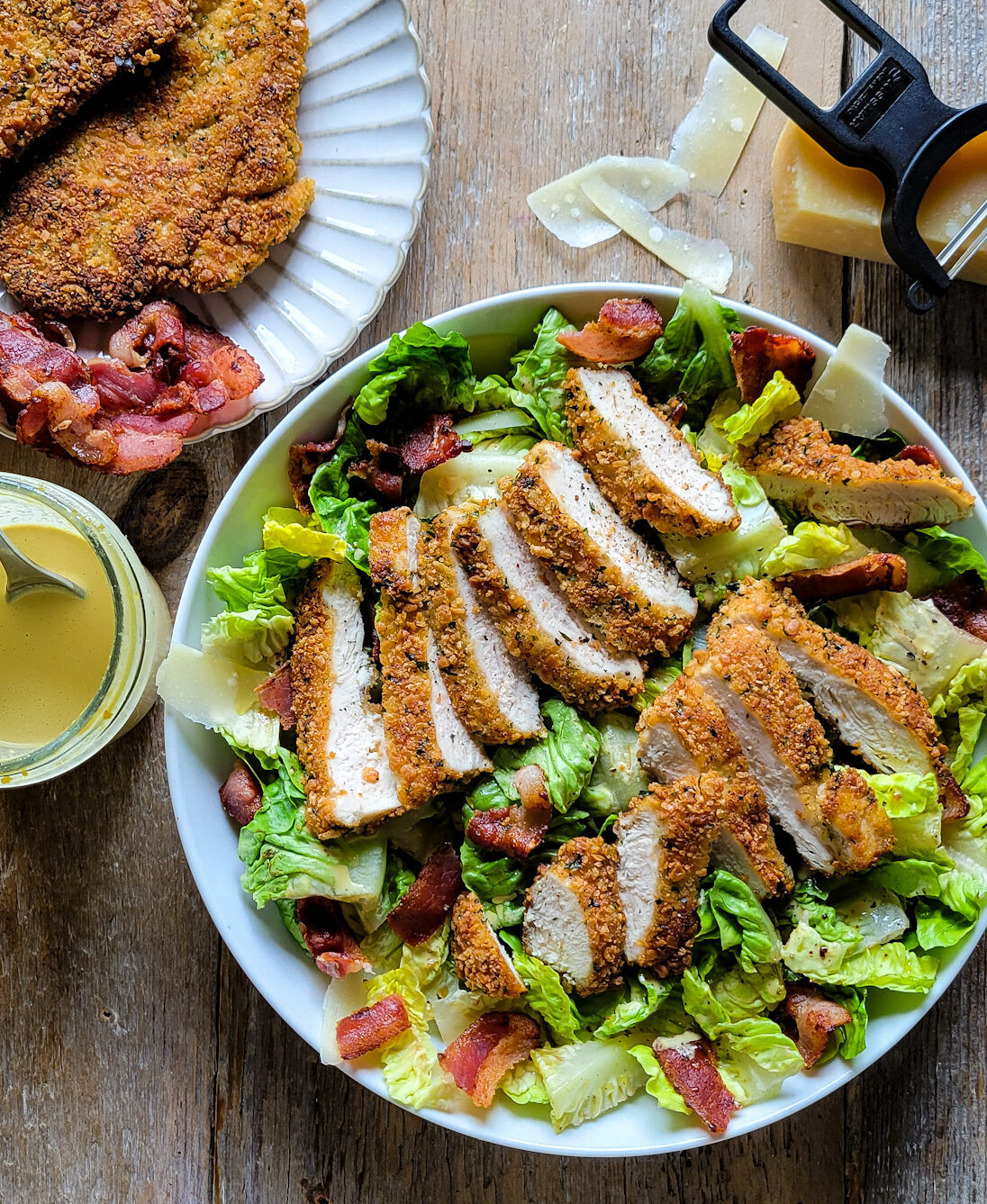 A bowl of Caesar Salad with sliced Parmesan Crusted Chicken on top with more chicken to the side as well as the creamy dressing and cheese.