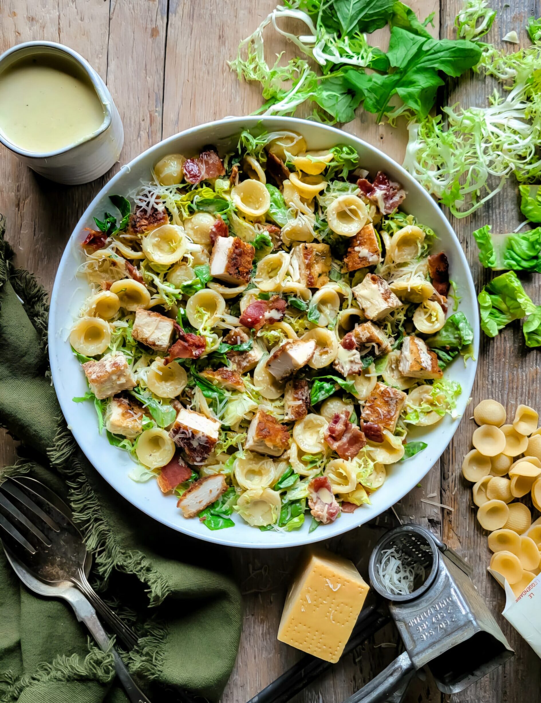 A bowl of Caesar Pasta Salad with Parmesan Crusted Chicken Croutons is surrounded by lettuce, cheese and creamy dressing.