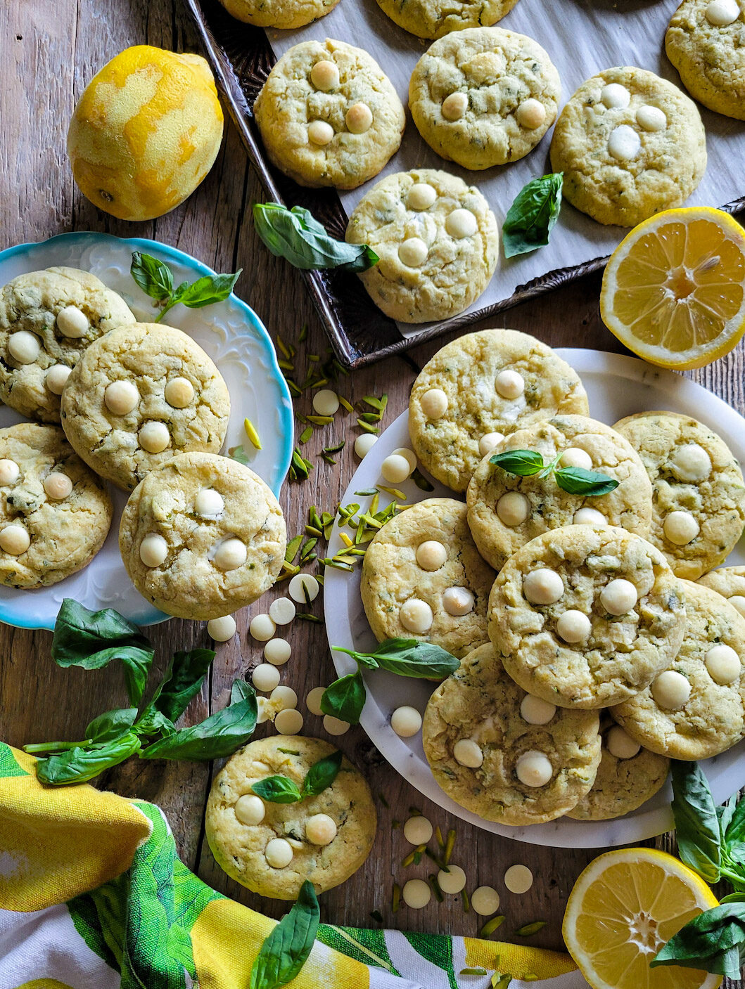 A sea of Lemon Basil Cookies with White Chocolate and Pistachios spread out on plates and baking sheet, surrounded by fresh basil leaves and sliced lemons.