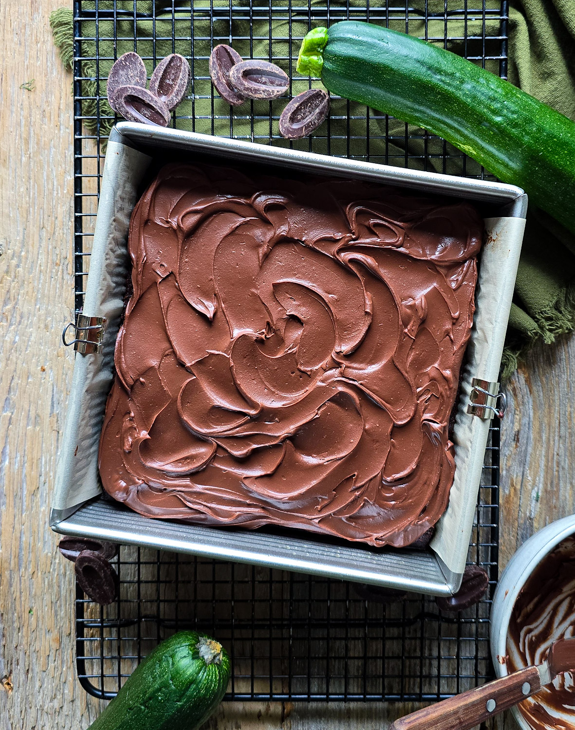 A Double Chocolate Zucchini Cake is frosted with Chocolate Ganache. The cake pan is surrounded by fresh zucchini and chocolate pieces.