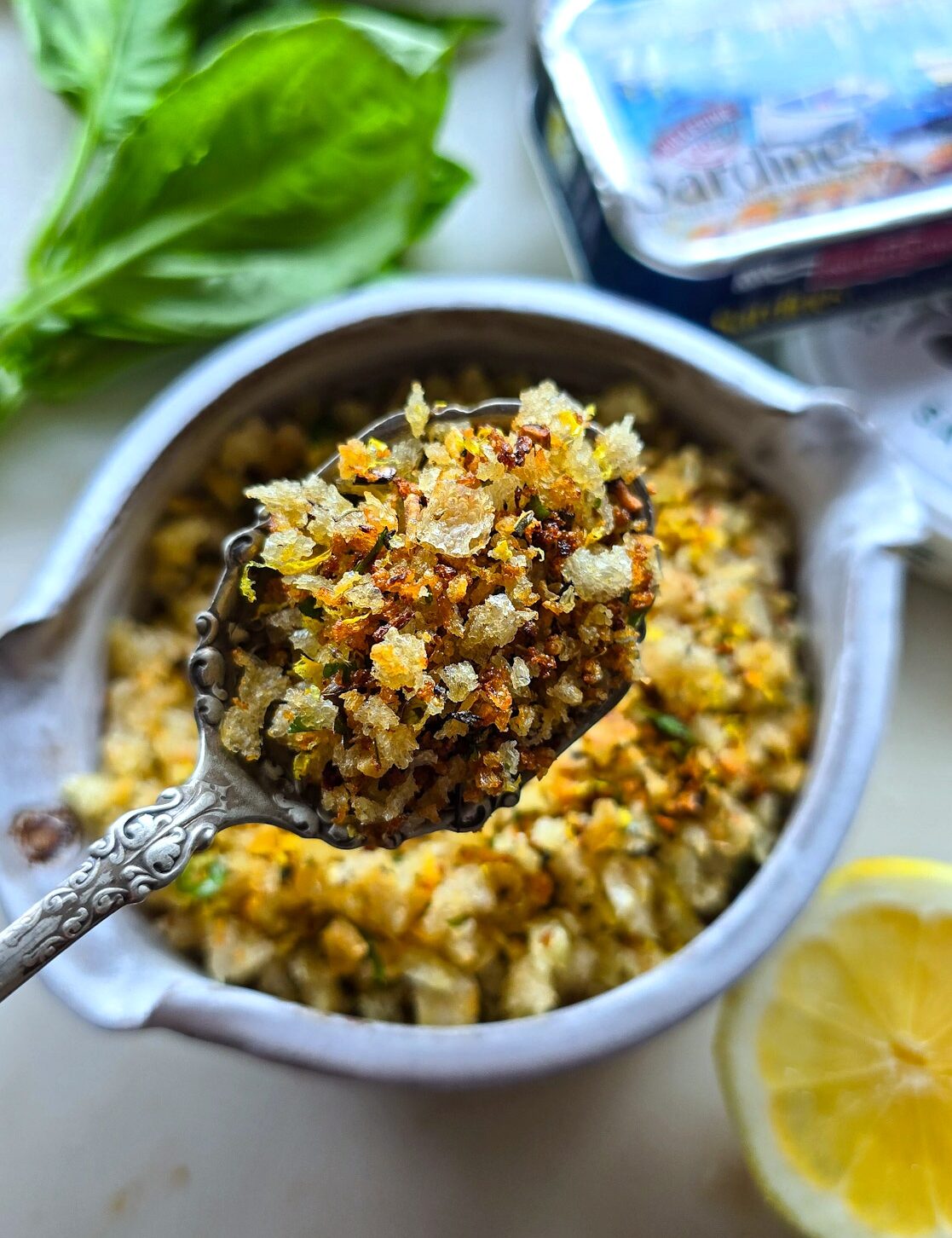 A bowl of crispy and bright Lemon Pangratttato on the counter, surrounded by a tin of sardines, and lemon slices.