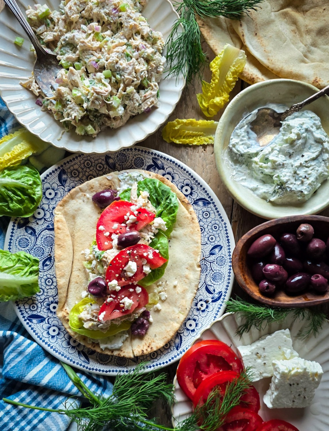 A table scape with all the ingredients needed to make a Greek Pita wrap using Tzatziki Chicken Salad. Olives, tomatoes and feta cheese surround the plate with the pita bread.