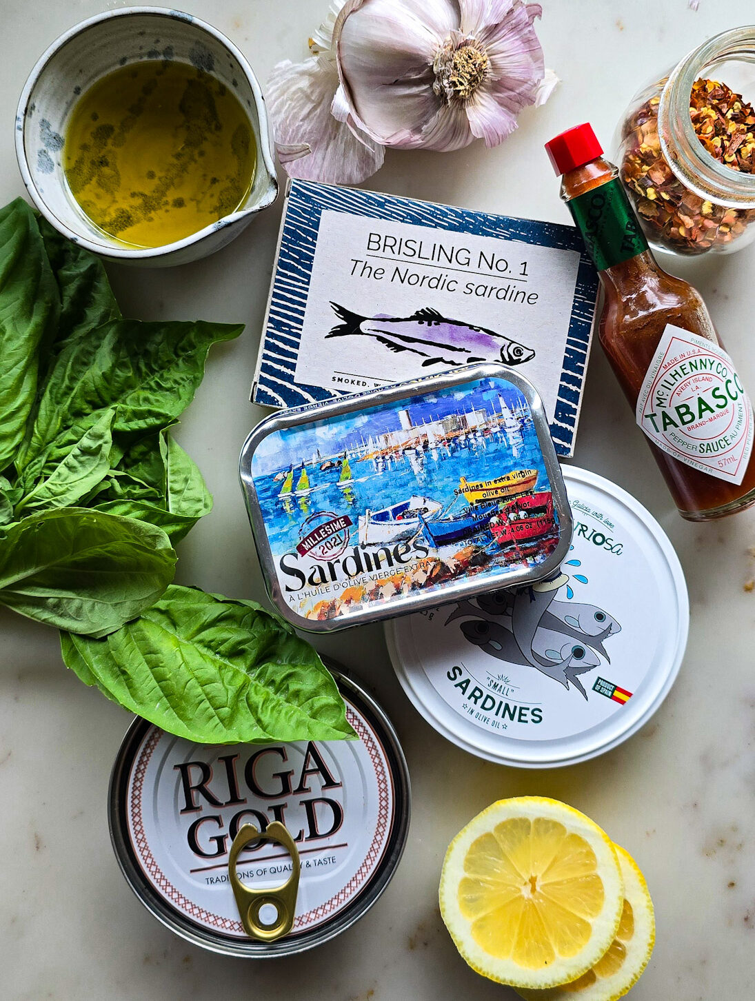 Various tins of fish like sardines and sprats are on the counter, with hot sauce, lemon, and basil leaves surrounding them.