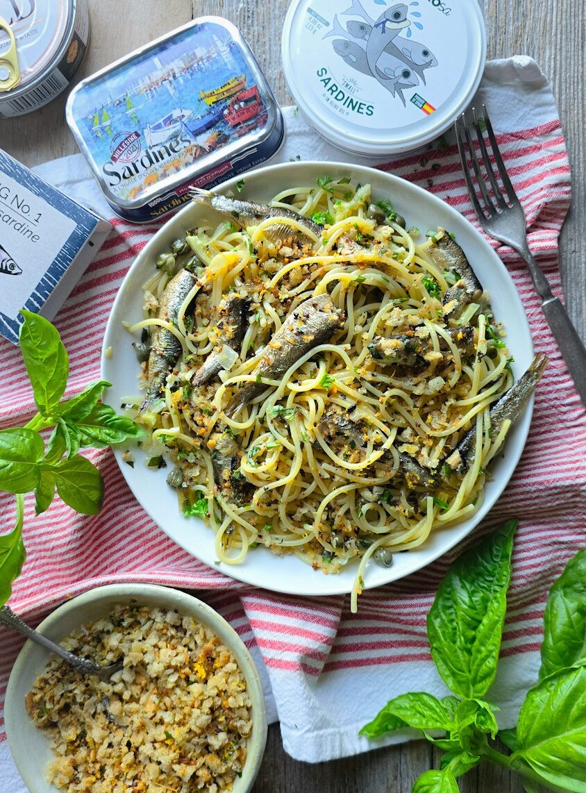 A plate of Sardine and Lemon Pangrattato Pasta sits on a tea towel, with tins of fish to the side, as well as a bowl of Lemon Pangrattato.