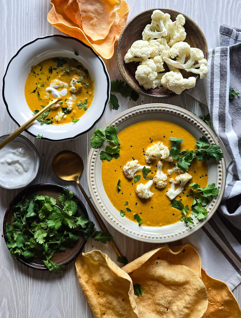 Two bowls of Curry Cauliflower Soup are on the table, with crispy pappadums on the side to eat with the soup.