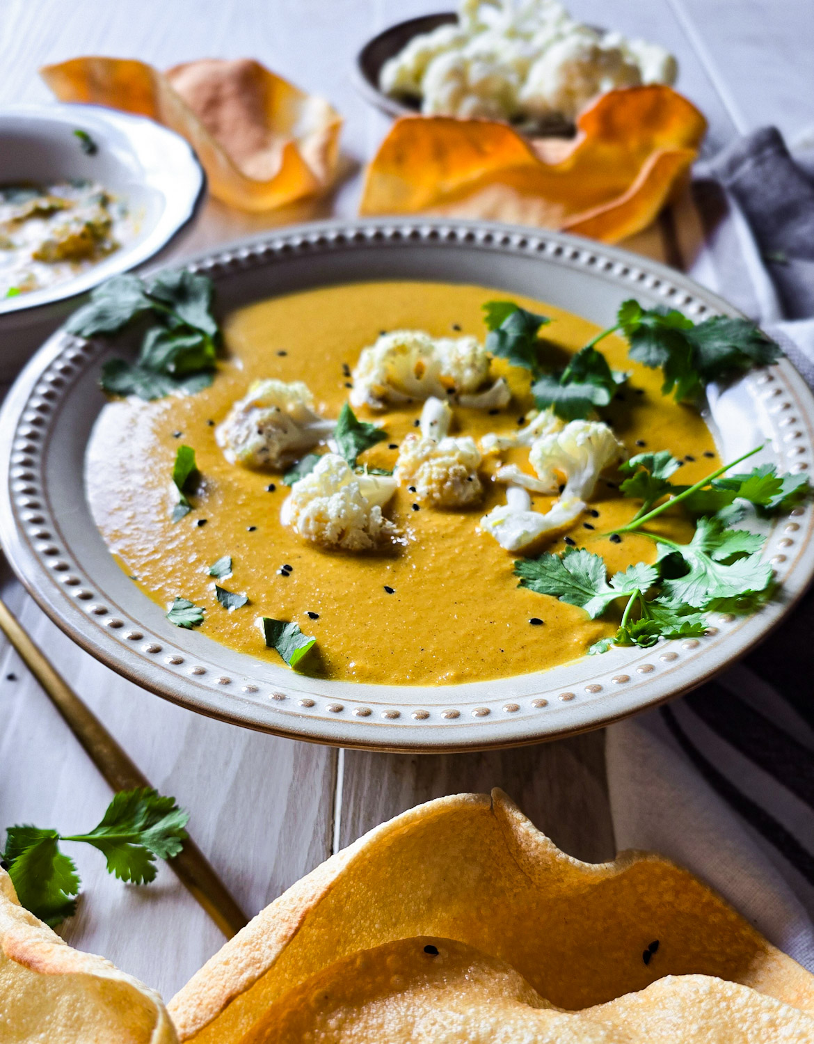 A bowl of Curry Cauliflower Soup with roasted cauliflower florets and cilantro scattered on top is on the table, with cripsy pappadums to the side.