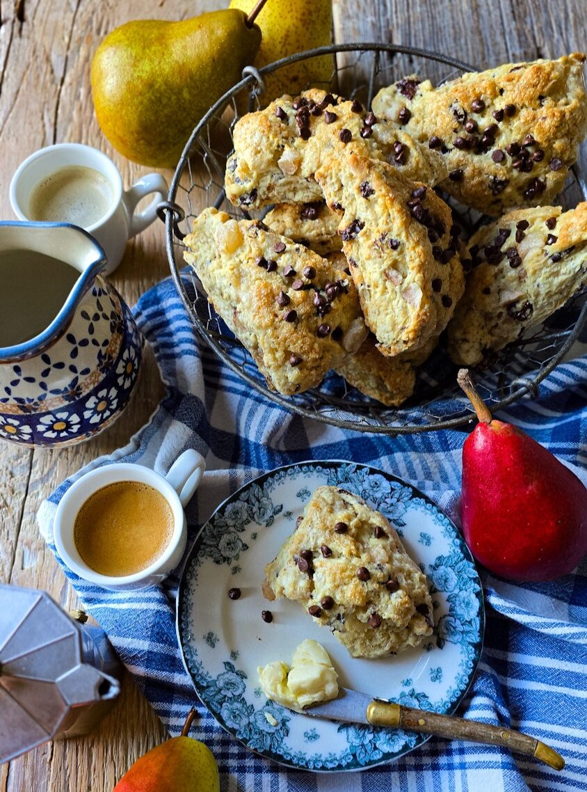 A Pear Chocolate Scone sits on a plate with a coffee to the side, and a basket of freshly baked scones in the background.