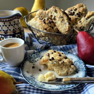 A Pear Chocolate Scone sits on a plate with a coffee to the side, and a basket of freshly baked scones in the background.