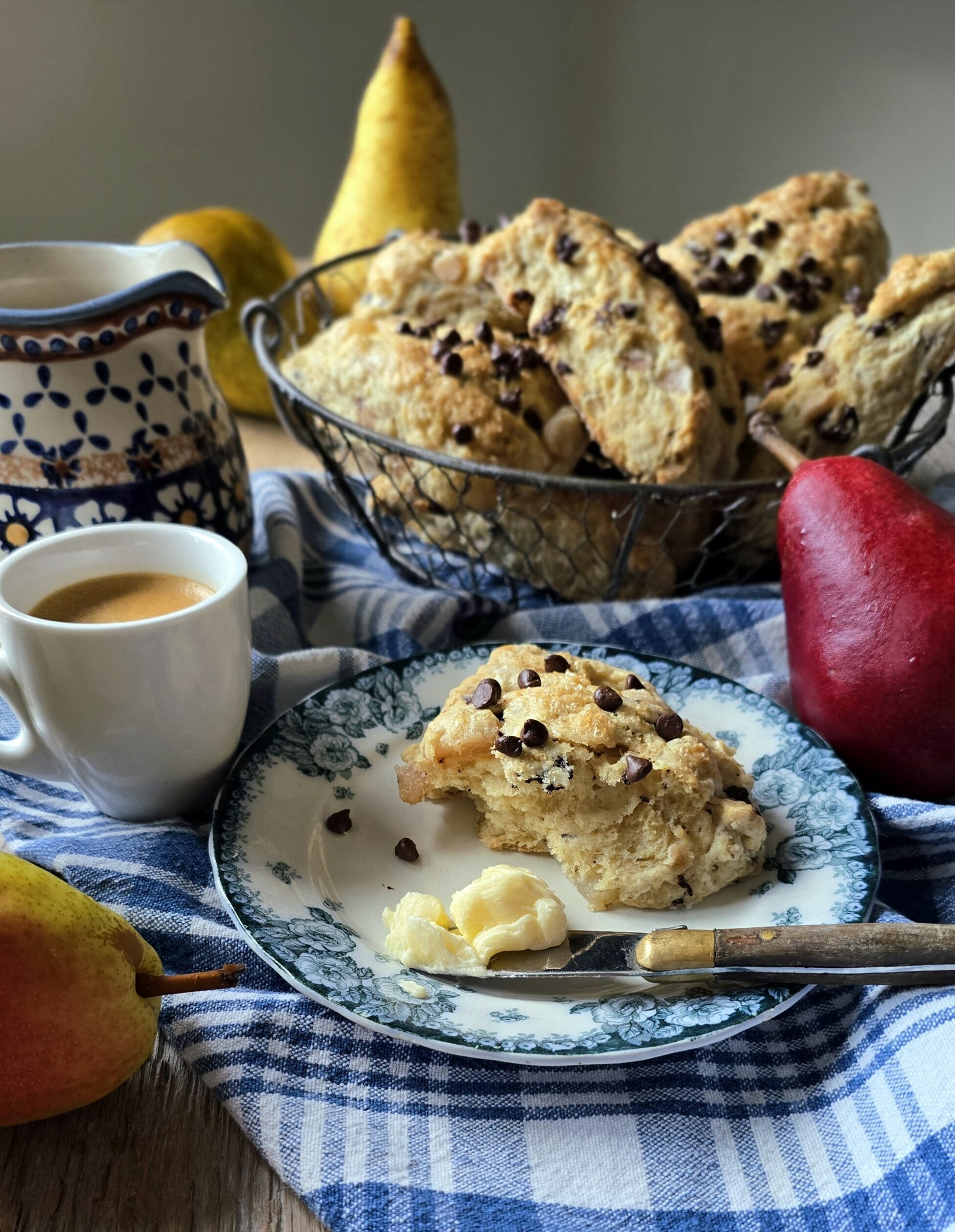 A Pear Chocolate Scone sits on a plate with a coffee to the side, and a basket of freshly baked scones in the background.