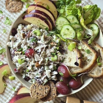 Close up of a Bowl filled with Leftover Diner Turkey Salad, with crackers, cucumbers, apple slices and grapes sitting on a table.