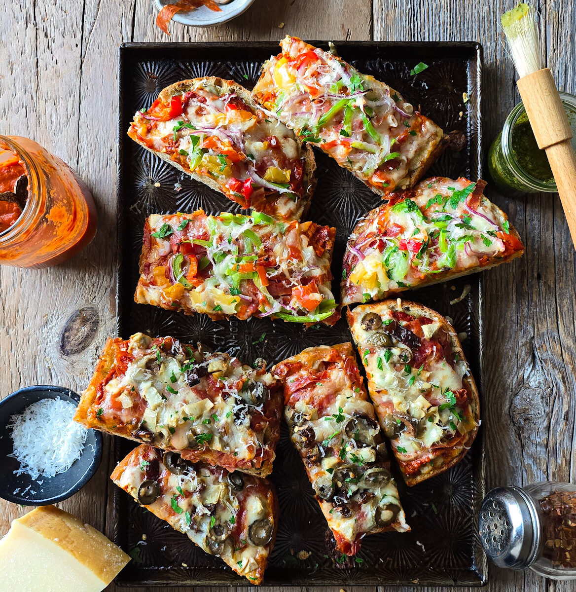 A baking sheet filled with French Bread Pizza is on the table, surrounded by homemade pizza sauce, parmesan cheese and pesto sauce.