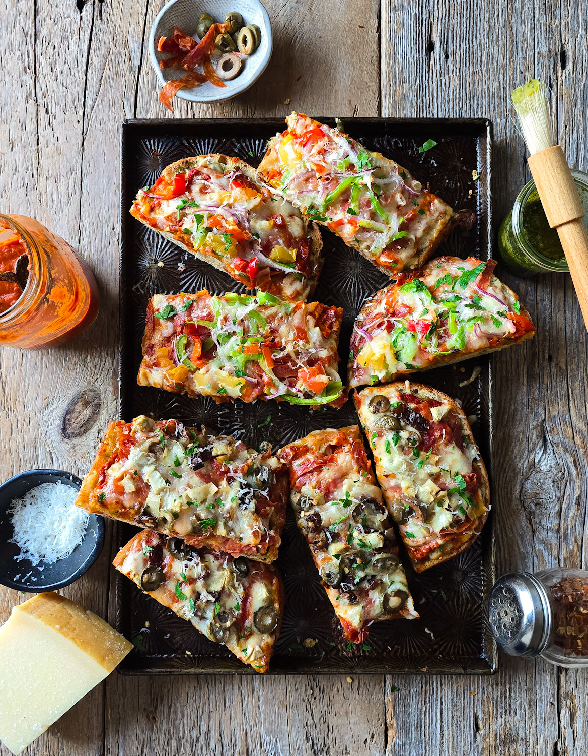 A baking sheet filled with French Bread Pizza is on the table, surrounded by homemade pizza sauce, parmesan cheese and pesto sauce.