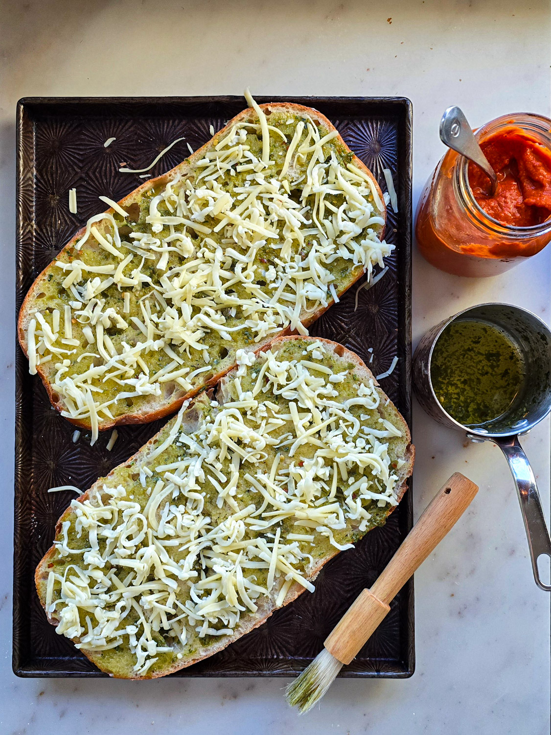 Pesto Garlic French Bread gets ready to be turned into Pizza. Two halves are sitting on the baking sheet ready to be baked off.