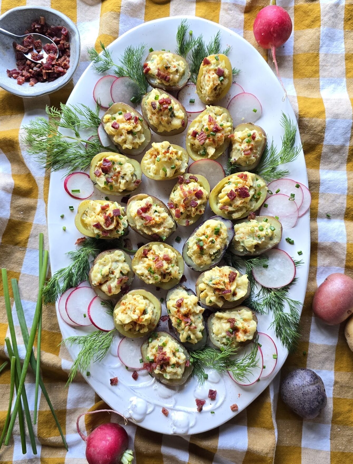 A platter filled with Devilled Mini Potatoes sits on the table surrounded by fresh dill fronds and a bowl of bacon bits