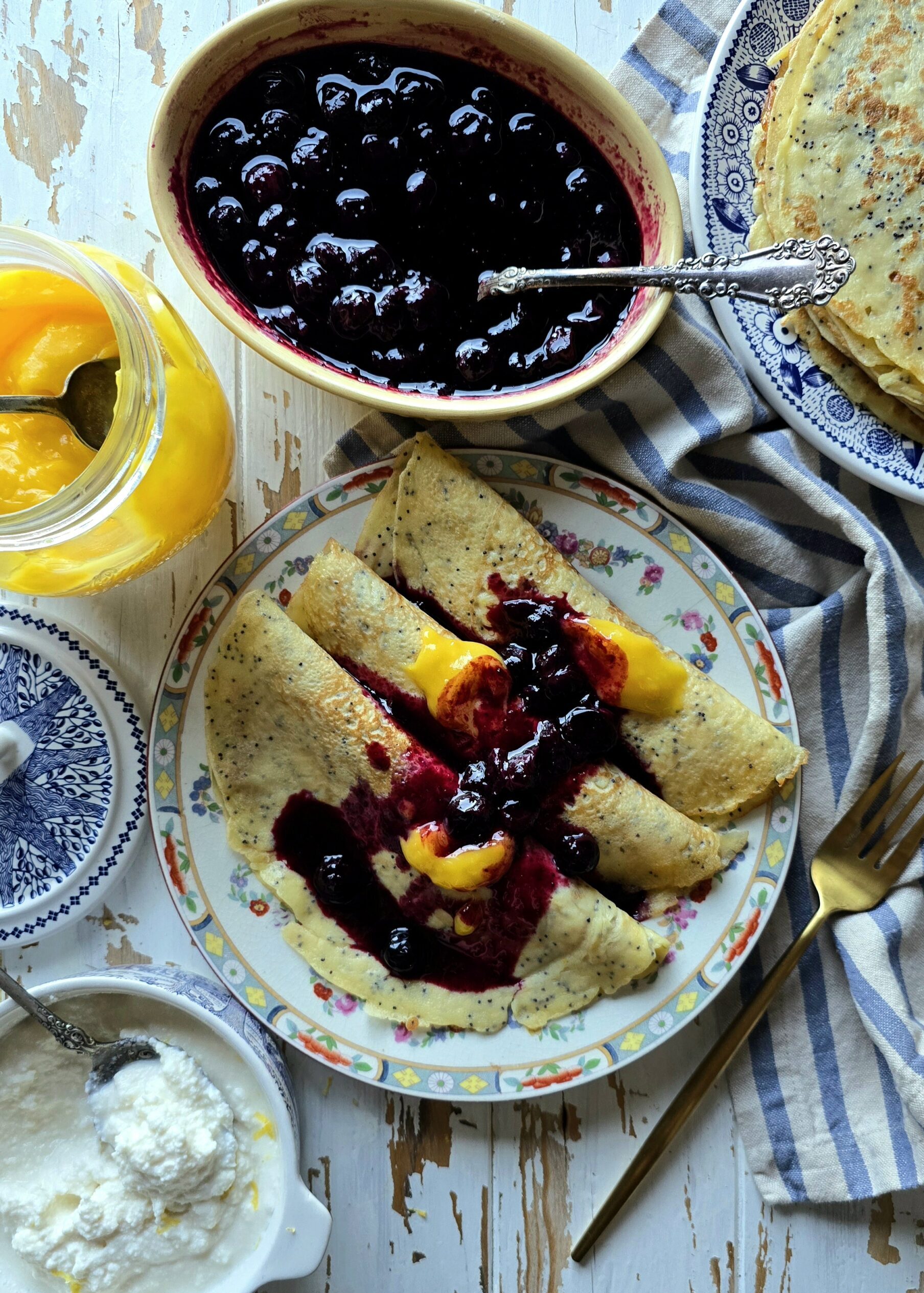 A plate of Lemon Poppyseed Crepes stuffed with sweet ricotta and lemon curd sits on the table, with Blueberry Sauce and more crepes to the side.