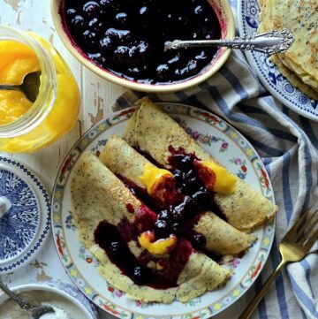 A plate of Lemon Poppyseed Crepes stuffed with sweet ricotta and lemon curd sits on the table, with Blueberry Sauce and more crepes to the side.