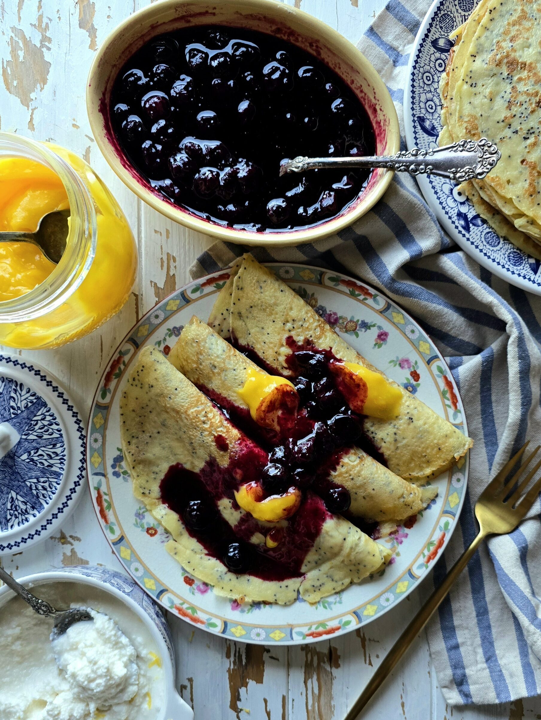 A plate of Lemon Poppyseed Crepes stuffed with sweet ricotta and lemon curd sits on the table, with Blueberry Sauce and more crepes to the side.
