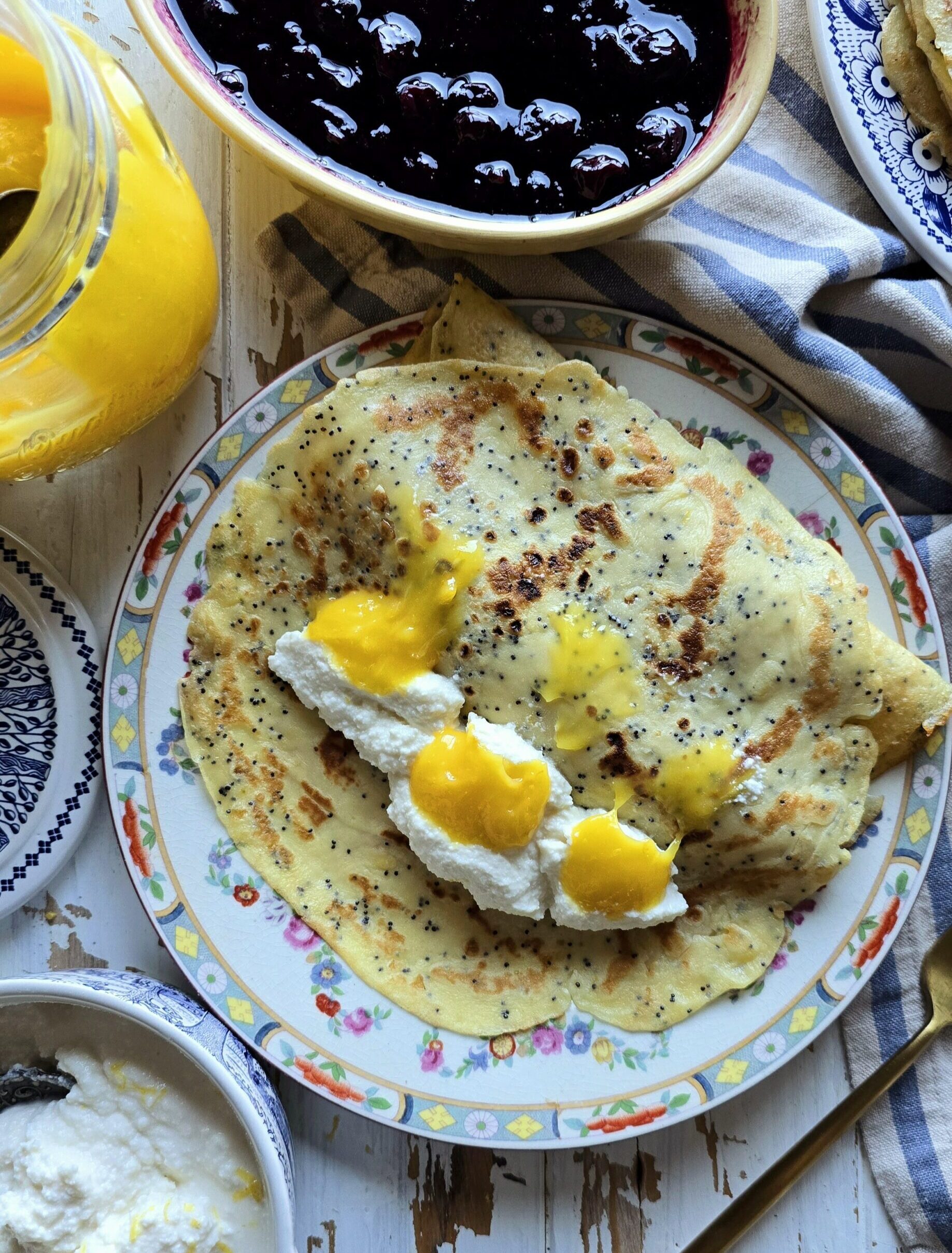A plate of Lemon Poppyseed Crepes shows the filling of sweet lemon ricotta and lemon curd. A bowl of Blueberry sauce is to the side.