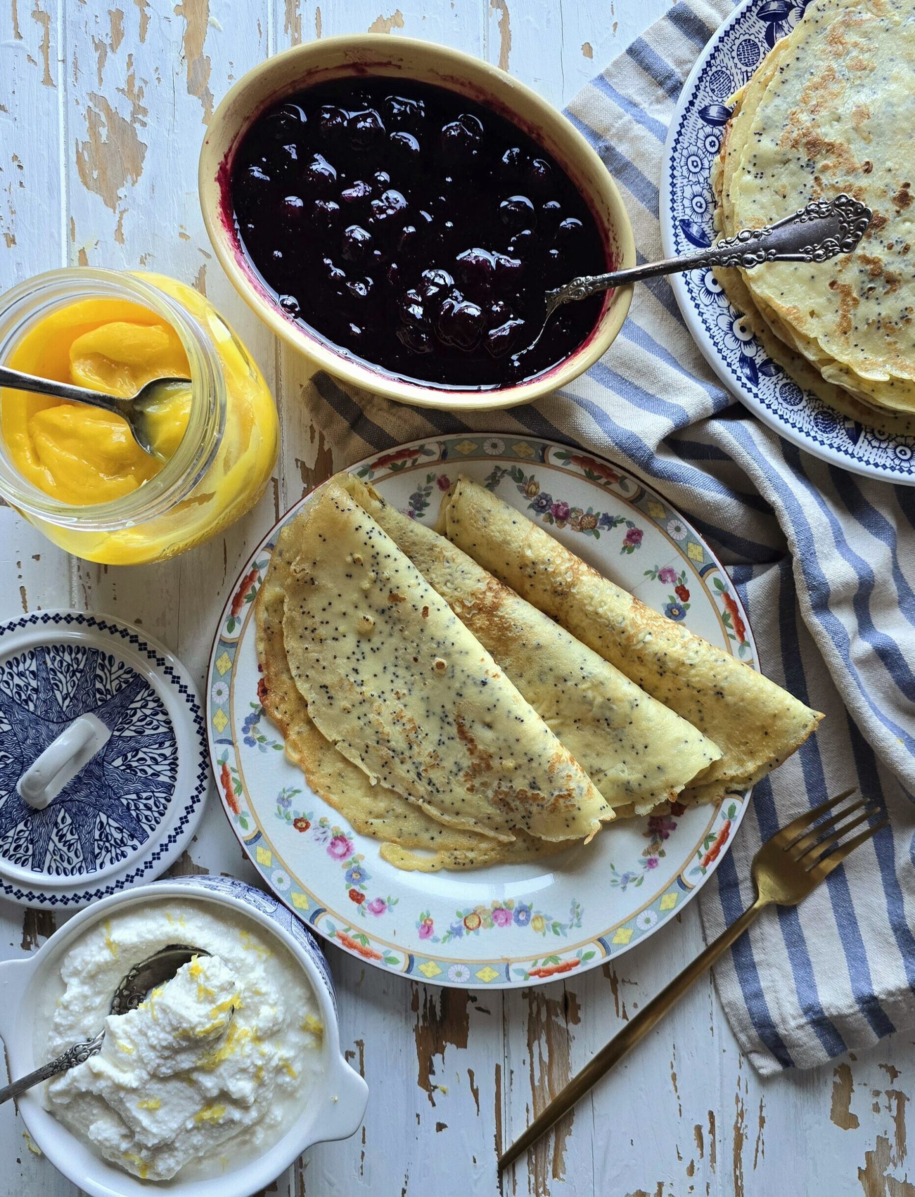 A table is set with Lemon Poppyseed Crepes, Blueberry Sauce, Lemon Curd and Sweet Lemon Ricotta ready to assemble the crepes.