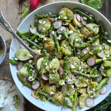 A serving bowl with Potato Asparagus Salad with Lemon Honey Vinaigrette is on the table, with a serving spoon and fresh radishes and dill to the side.