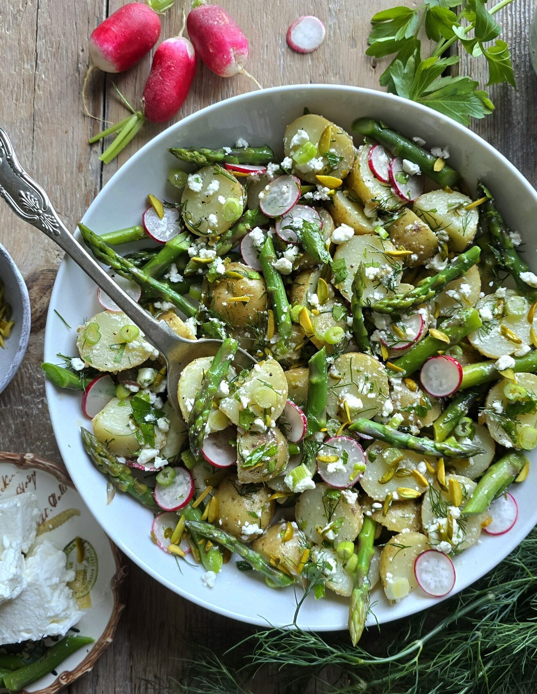 A serving bowl with Potato Asparagus Salad with Lemon Honey Vinaigrette is on the table, with a serving spoon and fresh radishes and dill to the side.