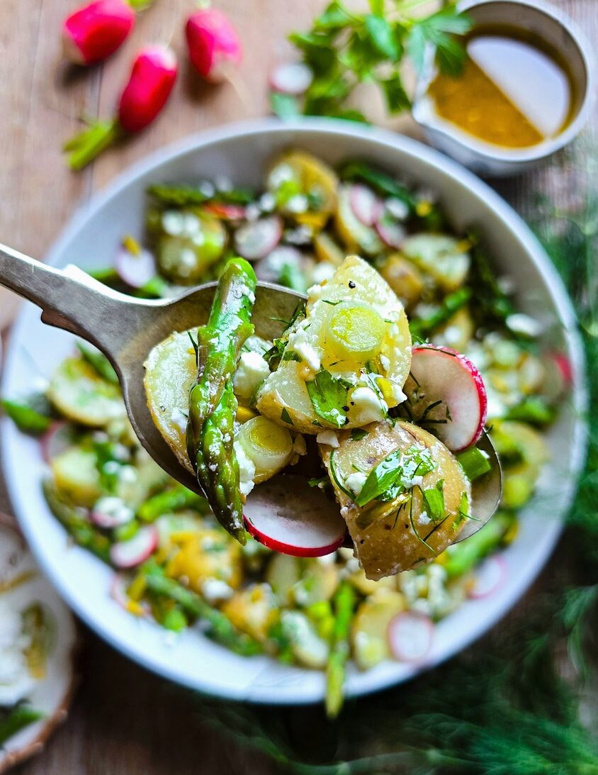 Close up of some Potato Asparagus Salad on a serving spoon above a bowl of the salad, with Lemon Honey Vinaigrette to the side.