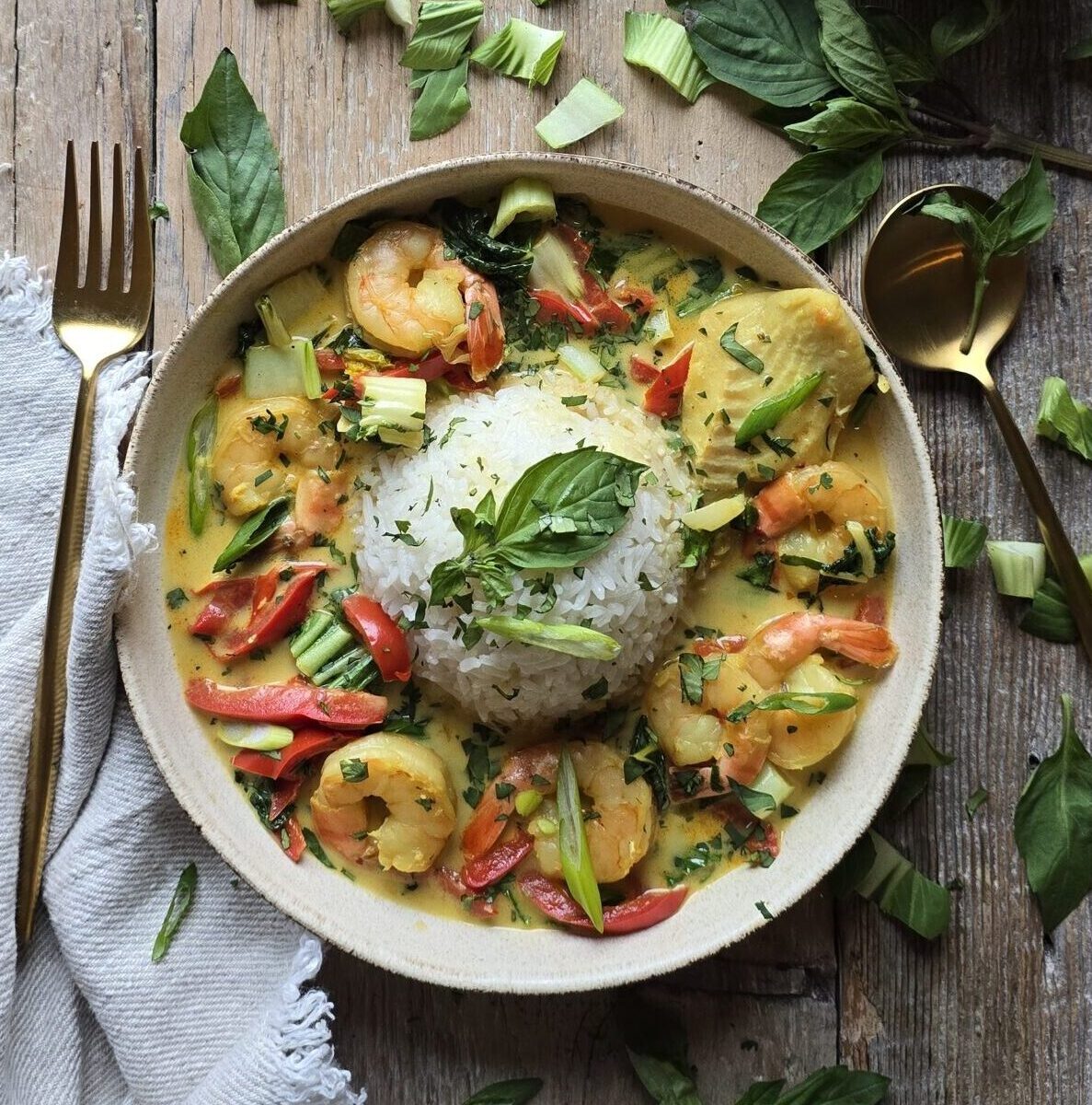 A bowl of Thai Fish Curry and rice sits on the table, with fresh cilantro and basil leaves to the side.