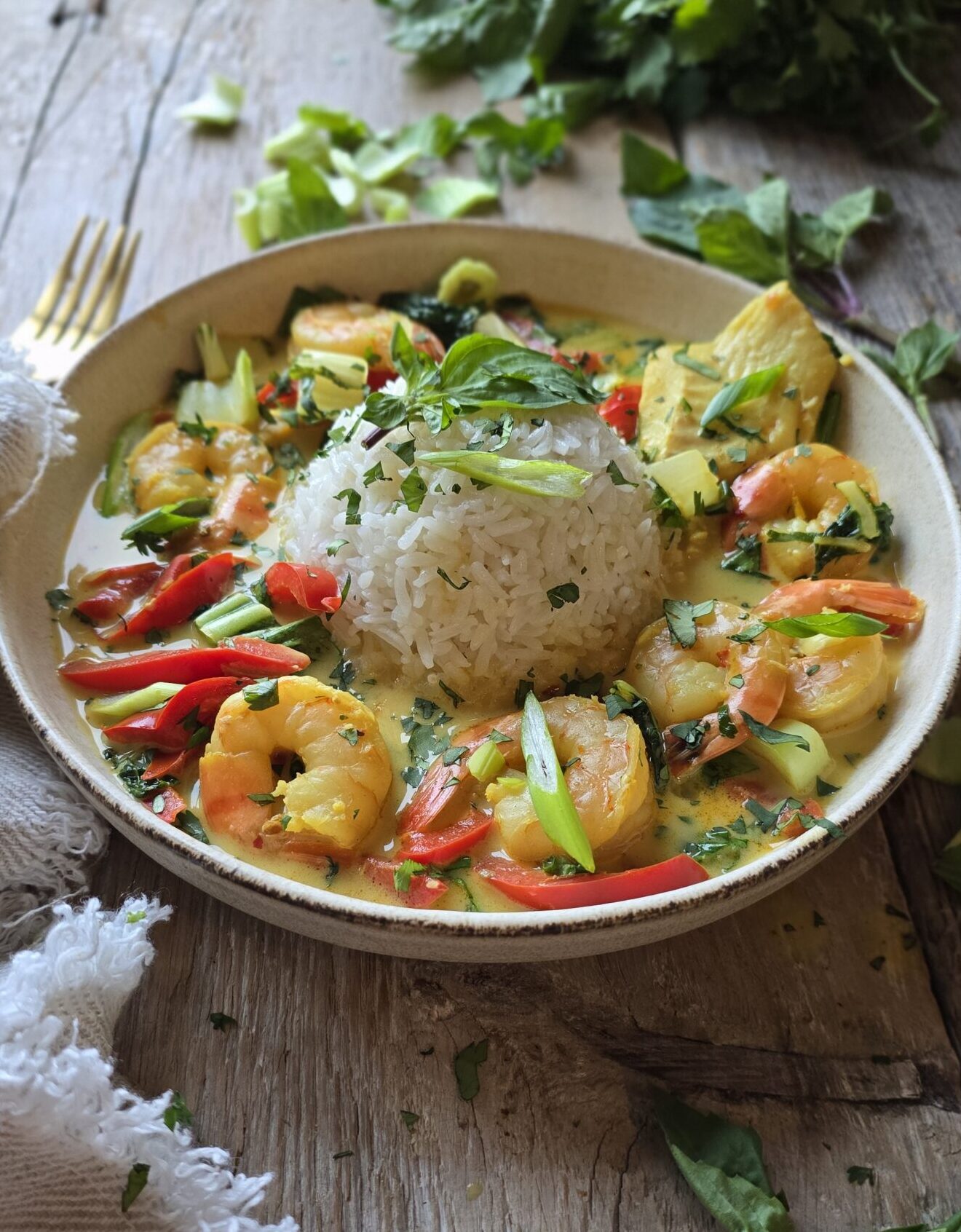 A bowl of Thai Fish Curry and rice sits on the table, surrounded by fresh cilantro and basil leaves.