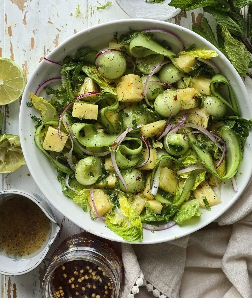A bowl of Pineapple Cucumber Salad with Mint sits on the table, with hot Honey vinaigrette to the side.