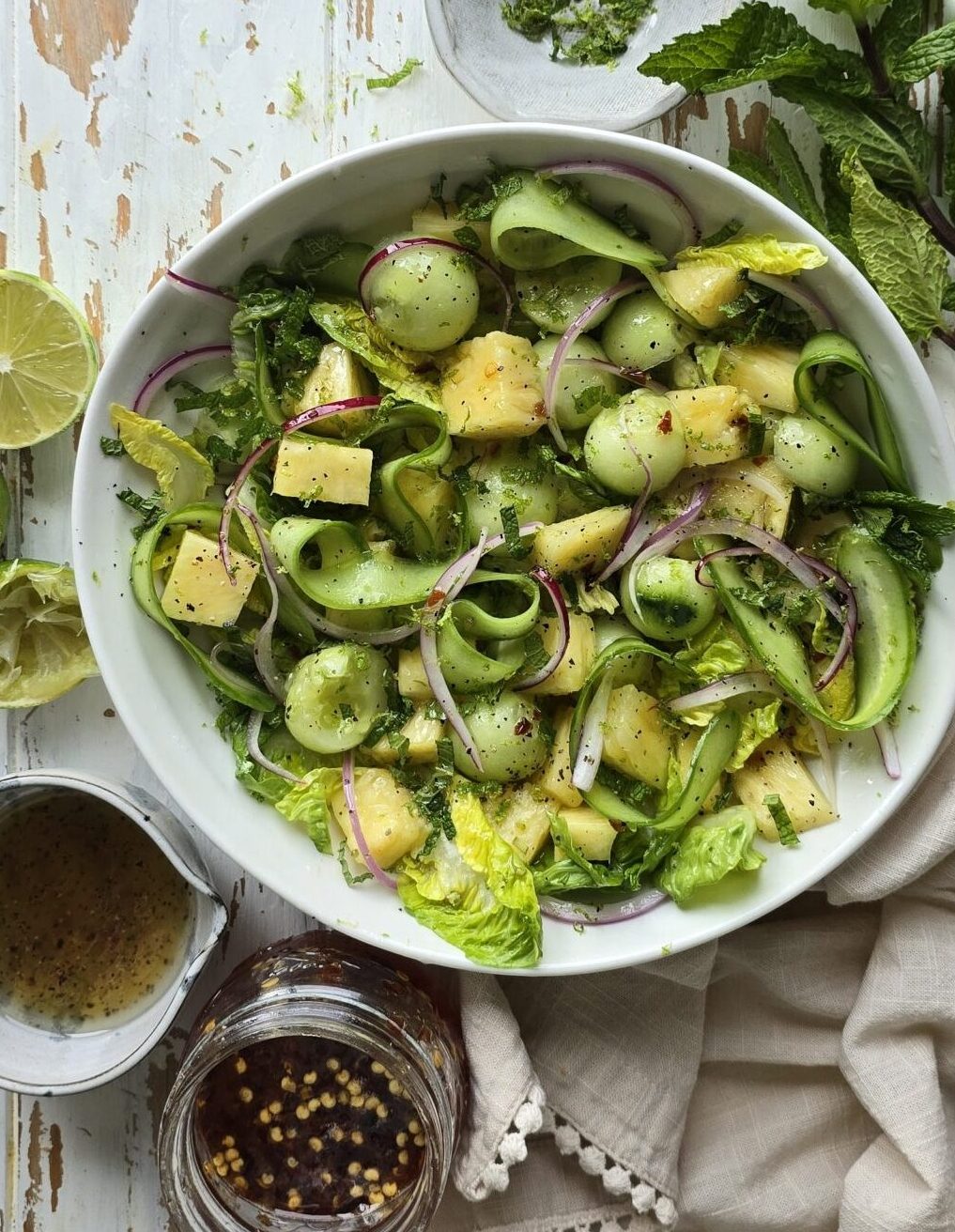 A bowl of Pineapple Cucumber Salad with Mint sits on the table, with hot Honey vinaigrette to the side.