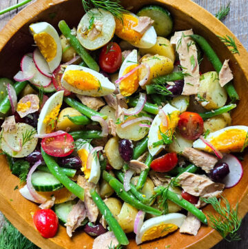 A wooden bowl is filled with a gently tossed Nicoise Potato Saald surrounded by fresh dill and parsley.