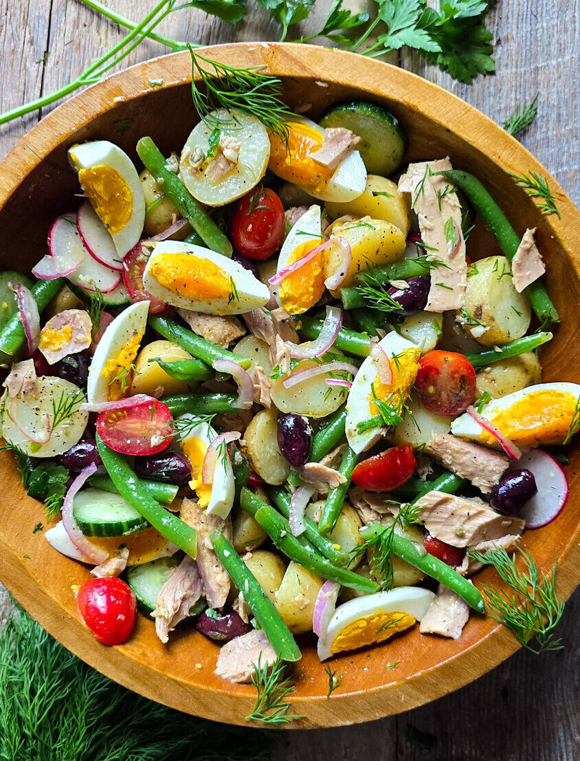 A wooden bowl is filled with a gently tossed Nicoise Potato Saald surrounded by fresh dill and parsley.