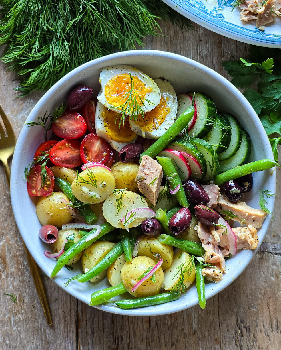 A Niçoise Potato Salad bowl is filled with all the fixings for a classic NIçoise Salad with fresh dill to the side.